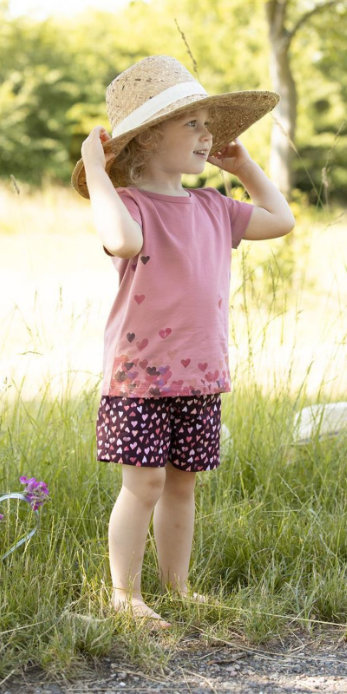 Mädchen im Sommeroutfit mit einem Sonnenhut, einem rosa Shirt mit Herzdruck von Enfant Terrible und eine dunkle Shorts mit hellen Herzen