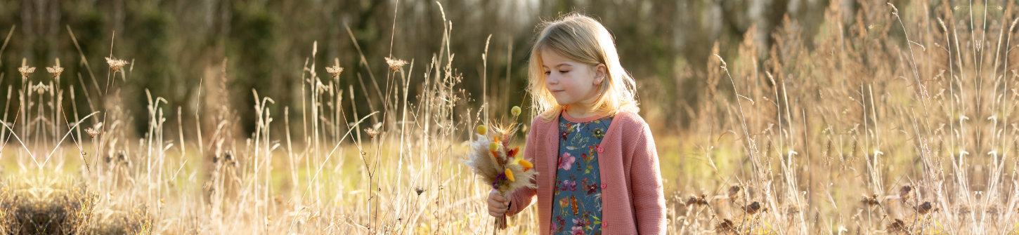 Ein fröhliches Mädchen steht im Freien und hält Blumen. Sie trägt nachhaltige Kindermode aus dem Naturladen in Braunschweig.